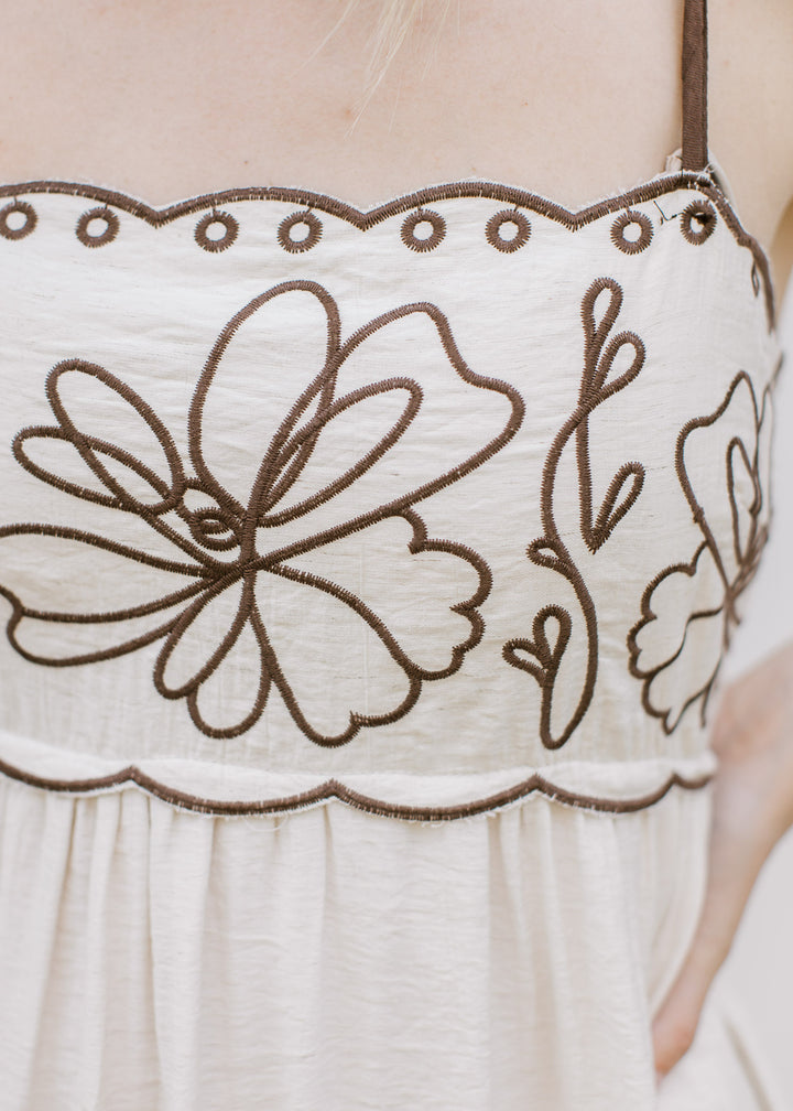 Close up of scalloped detail, brown embroidered floral and spaghetti straps on a cream midi.