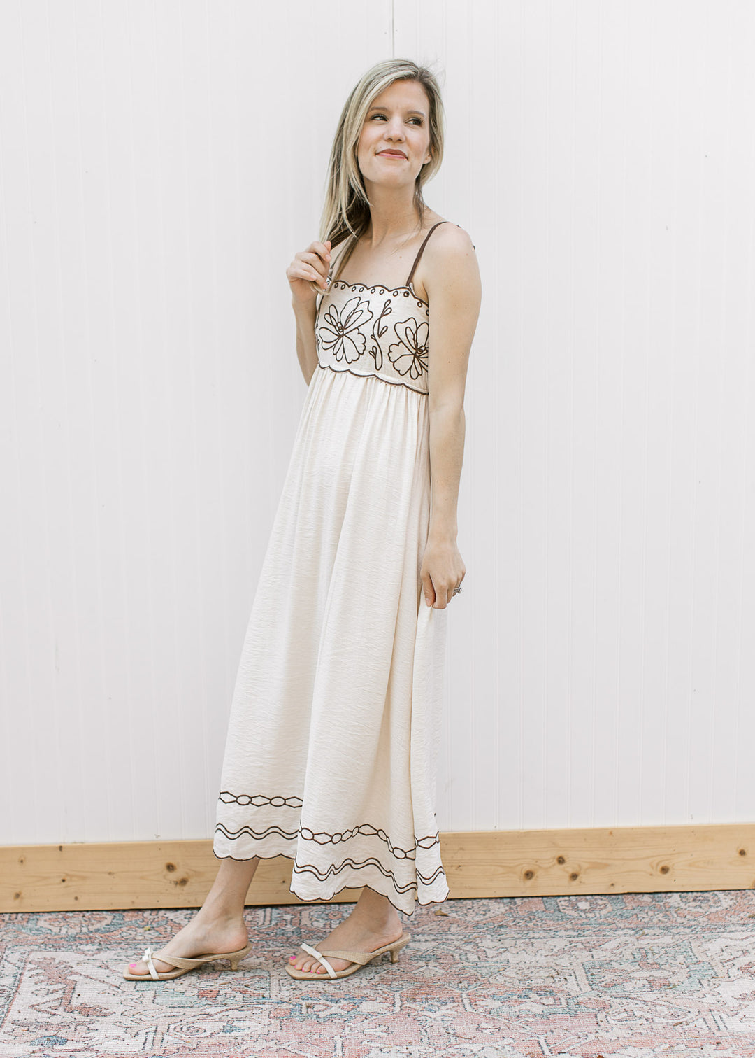 Model wearing sandals and a cream spaghetti strap midi with brown embroidery. 