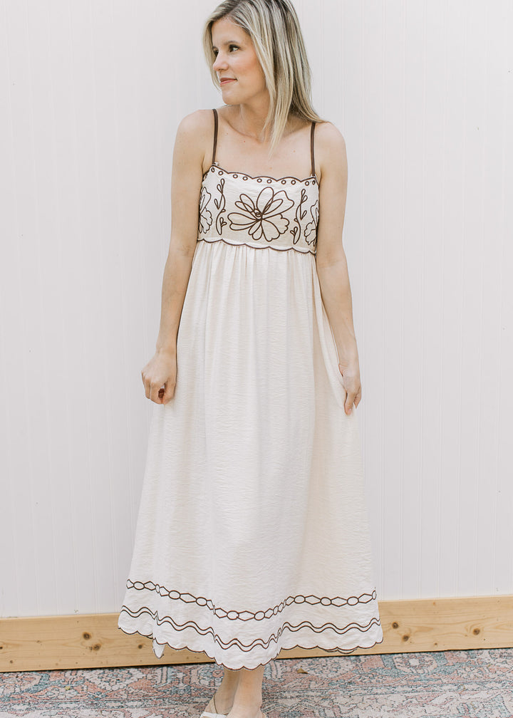 Model wearing a cream spaghetti strap midi with brown embroidered floral and a square neck. 