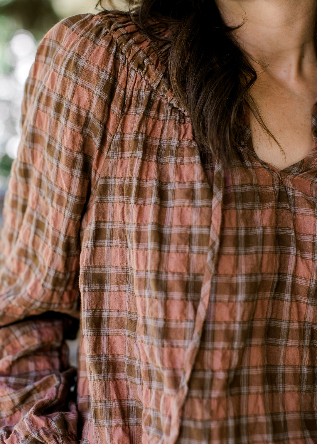 Close up of smocked neckline and v-neck with a tie on a blush and brown gingham top. 