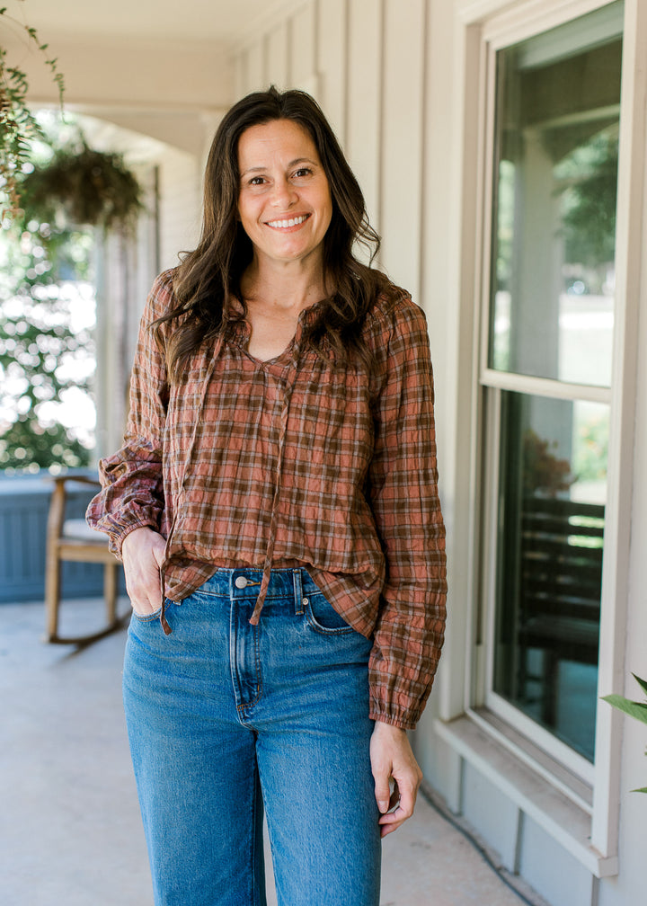 Model wearing jeans and a long sleeve blush and brown gingham top with a smocked neck.