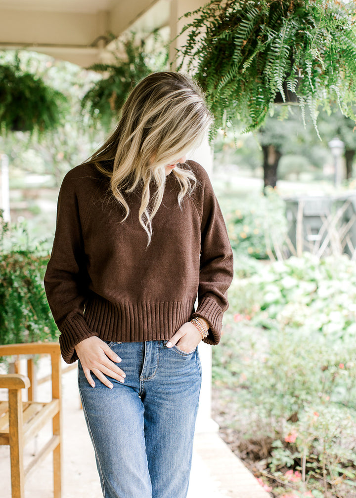 Model wearing jeans and a brown sweater with a mock neckline and long sleeves with ribbing detail.