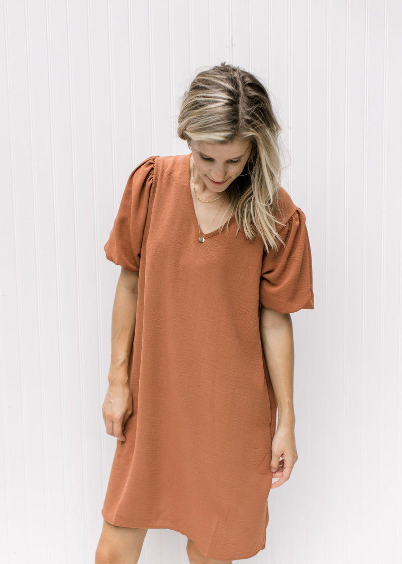 Model wearing a rust colored above the knee dress with bubble short sleeves and pockets. 