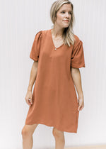 Model wearing a rust colored, v-neck dress with bubble short sleeves and pockets. 