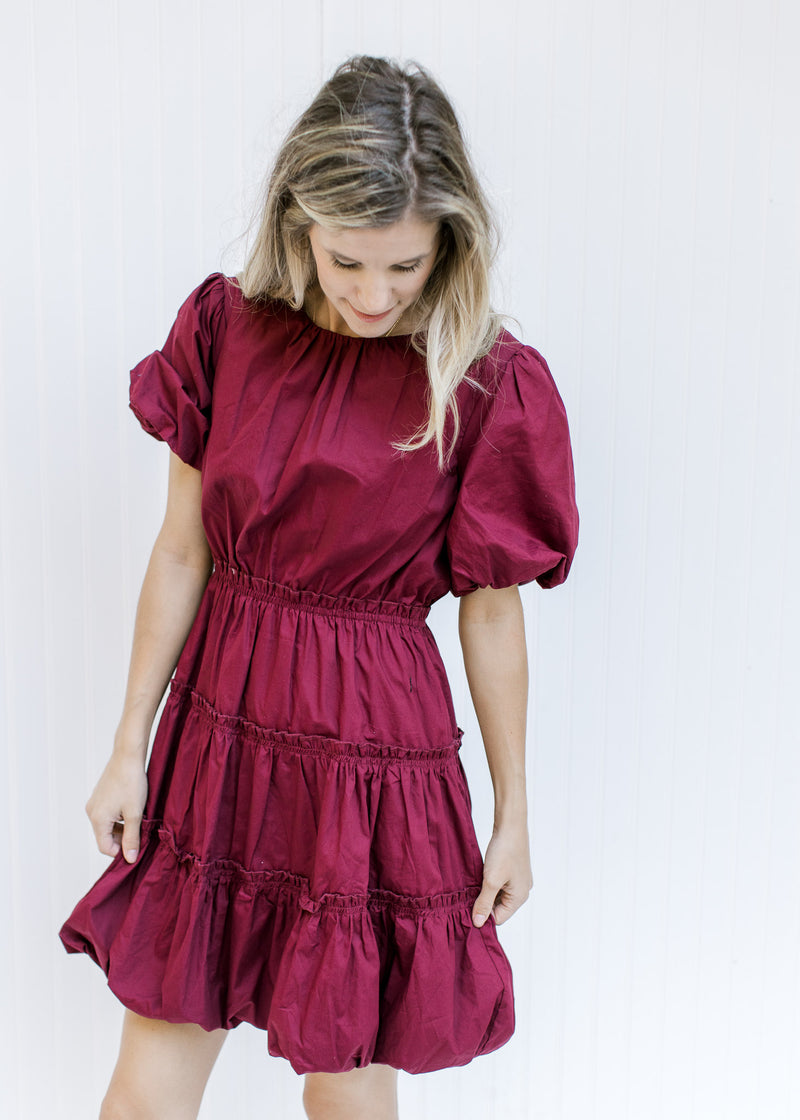Model wearing a burgundy above the knee dress with bubble short sleeves and hem and a round neck.