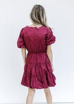 Back view of a Model wearing a burgundy above the knee dress with bubble short sleeves and tiers. 