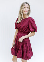 Model wearing a burgundy above the knee dress with tiers and bubble short sleeves and hem. 