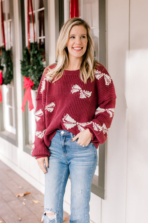 Model wearing a burgundy long sleeve sweater with cream bows that are embellished with pearl beads. 