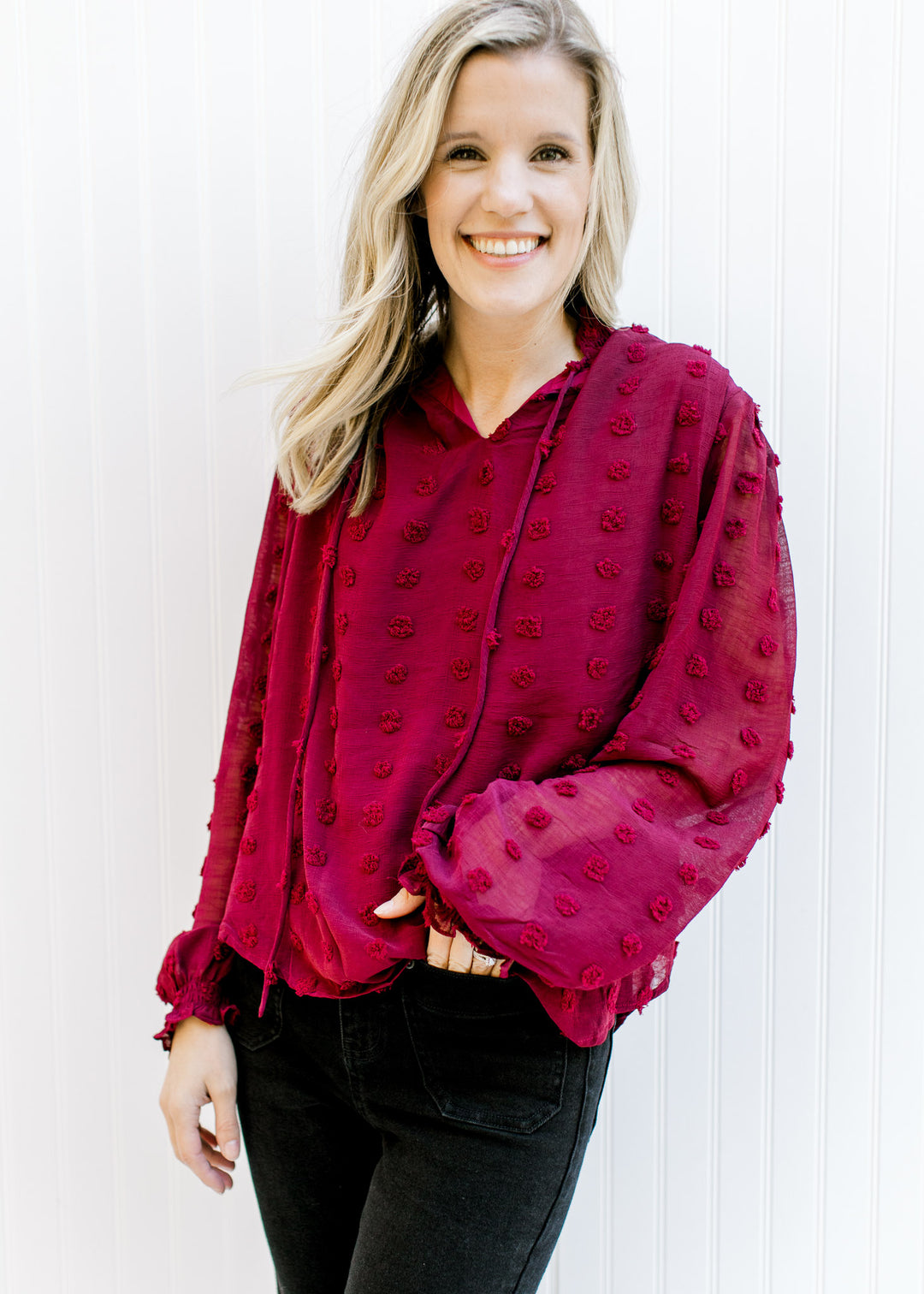 Model wearing a wine dotted Swiss top with sheer sleeves, a mock neckline and a tie at the neck. 