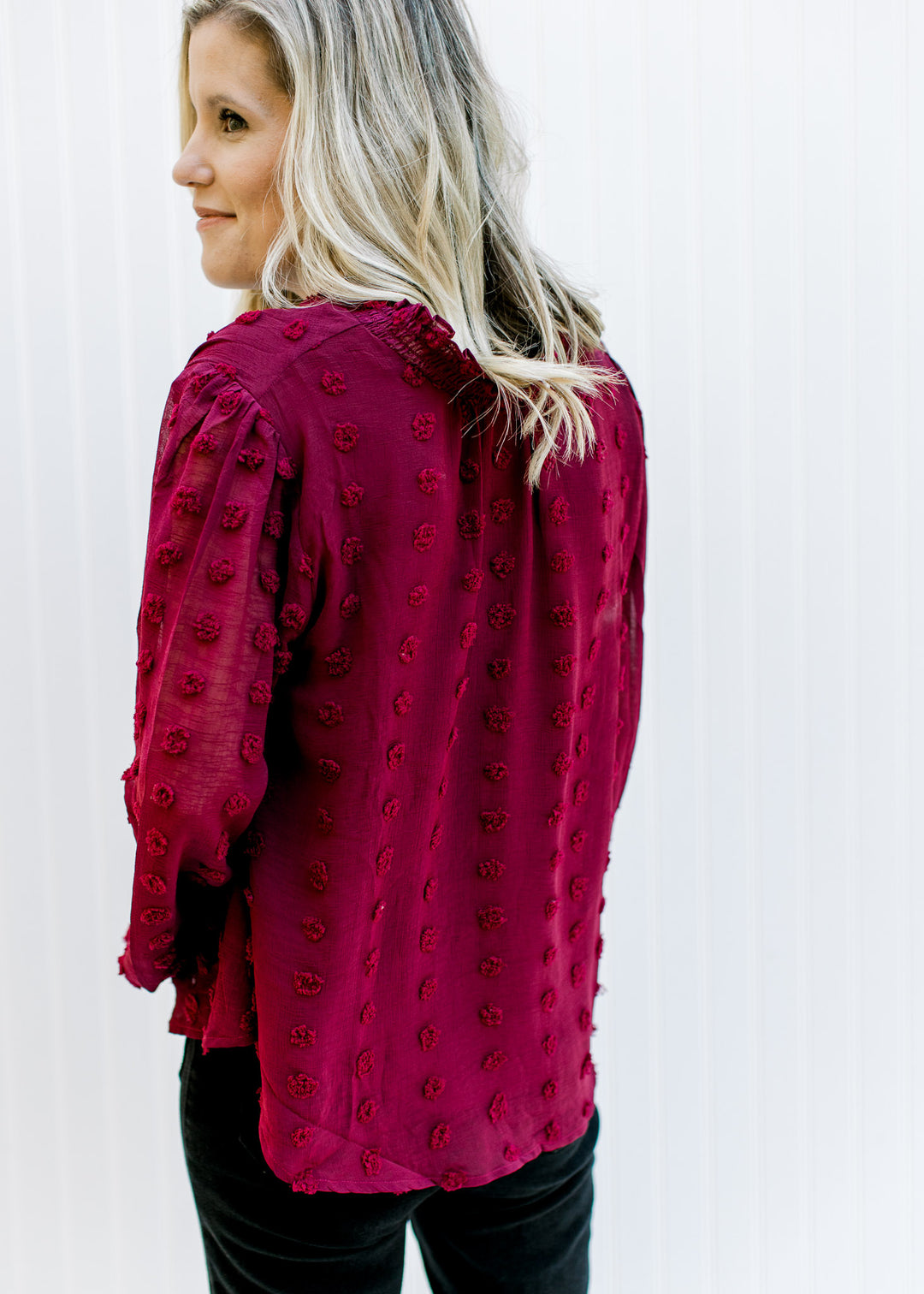 Back view of Model wearing a wine dotted Swiss top with sheer sleeves and mock neck with a tie. 