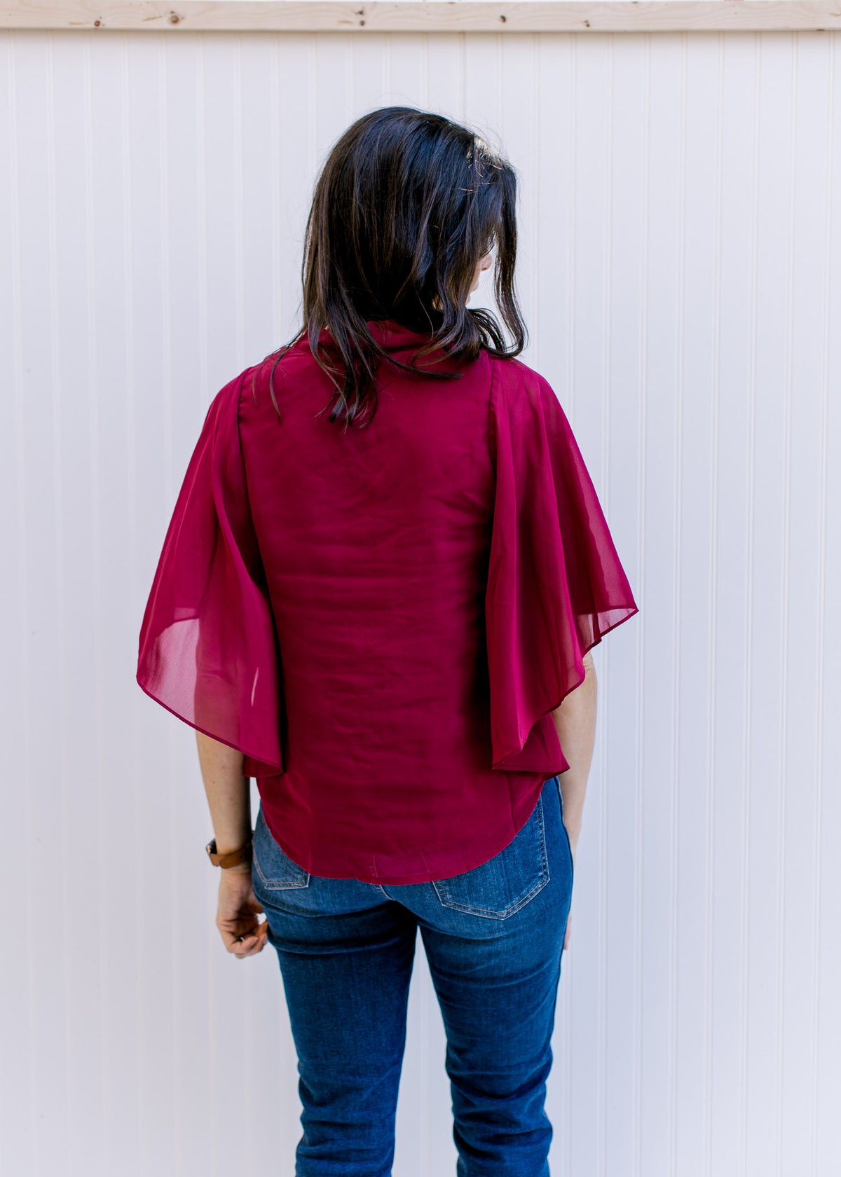 Back view of Model wearing a fully lined, burgundy blouse with kimono sleeves and v-neck. 