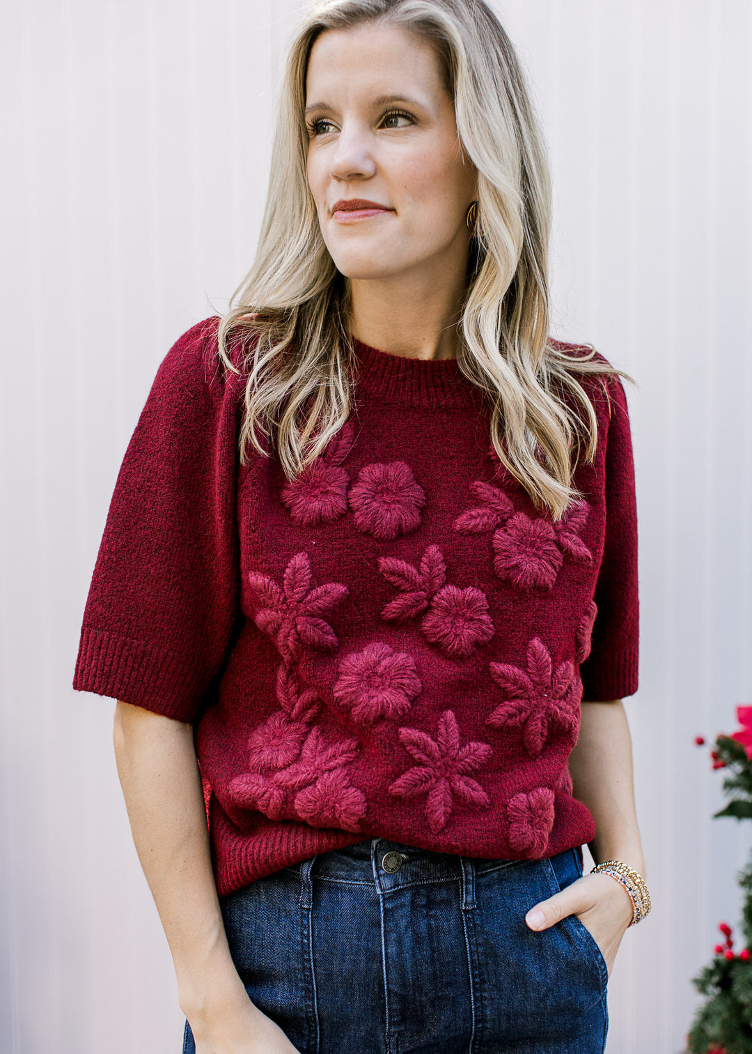 Model wearing a short sleeve burgundy sweater with 3D flowers and a round neck with ribbed detail.