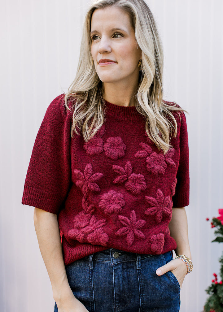 Model wearing a short sleeve burgundy sweater with 3D flowers and a round neck with ribbed detail.