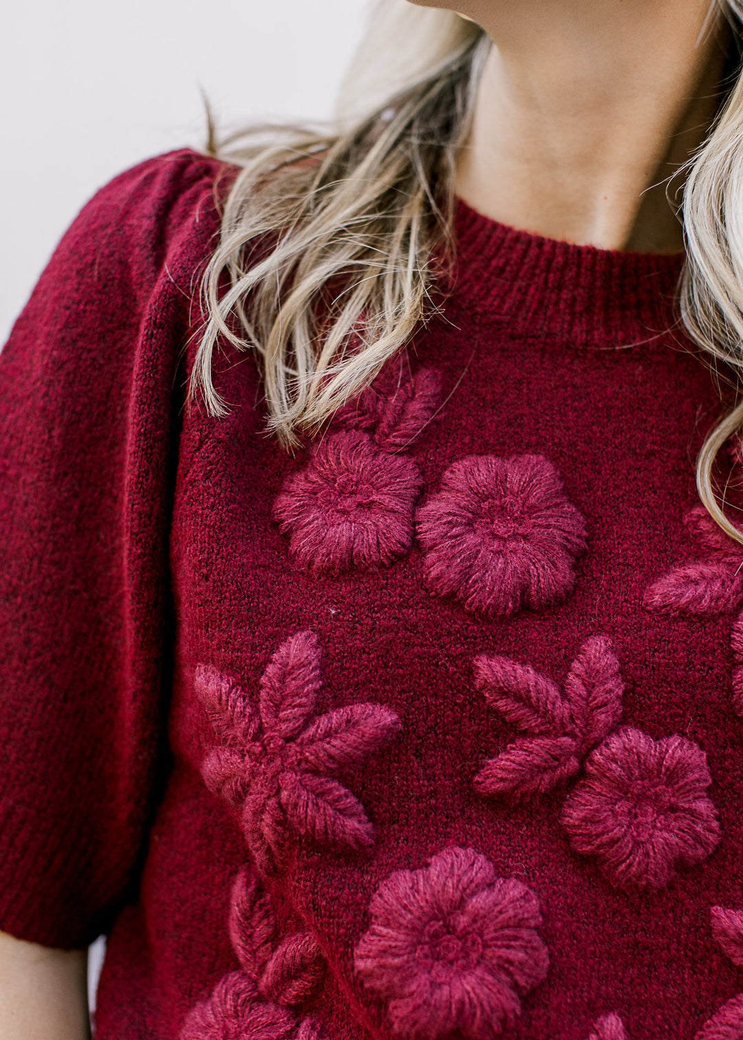 Model wearing a burgundy sweater with stitched 3D flowers, short sleeves and a round neckline. 
