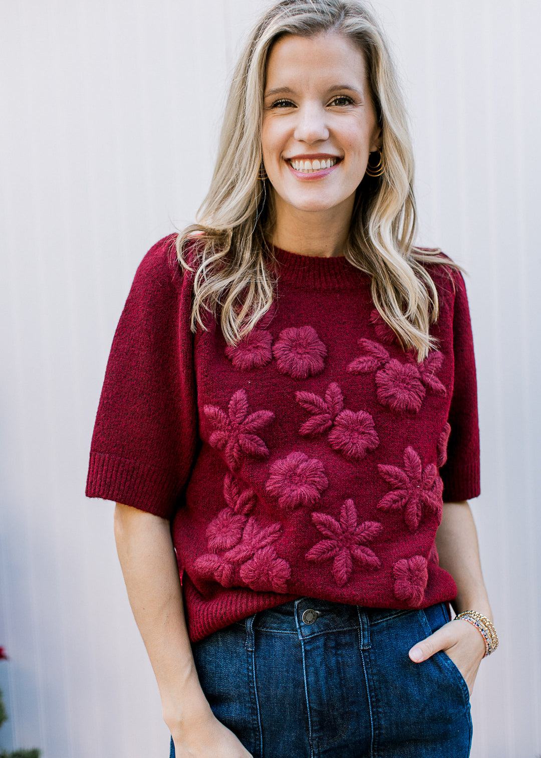 Model wearing a burgundy sweater with 3D flowers across the front, short sleeves and a round neck. 