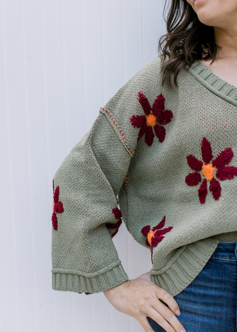 Close up view of burgundy flowers with an orange center on a sage long sleeve sweater with a v-neck.