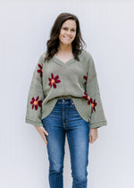 Model wearing jeans and a sage sweater with burgundy flowers, exposed hem, long sleeves and  v-neck.