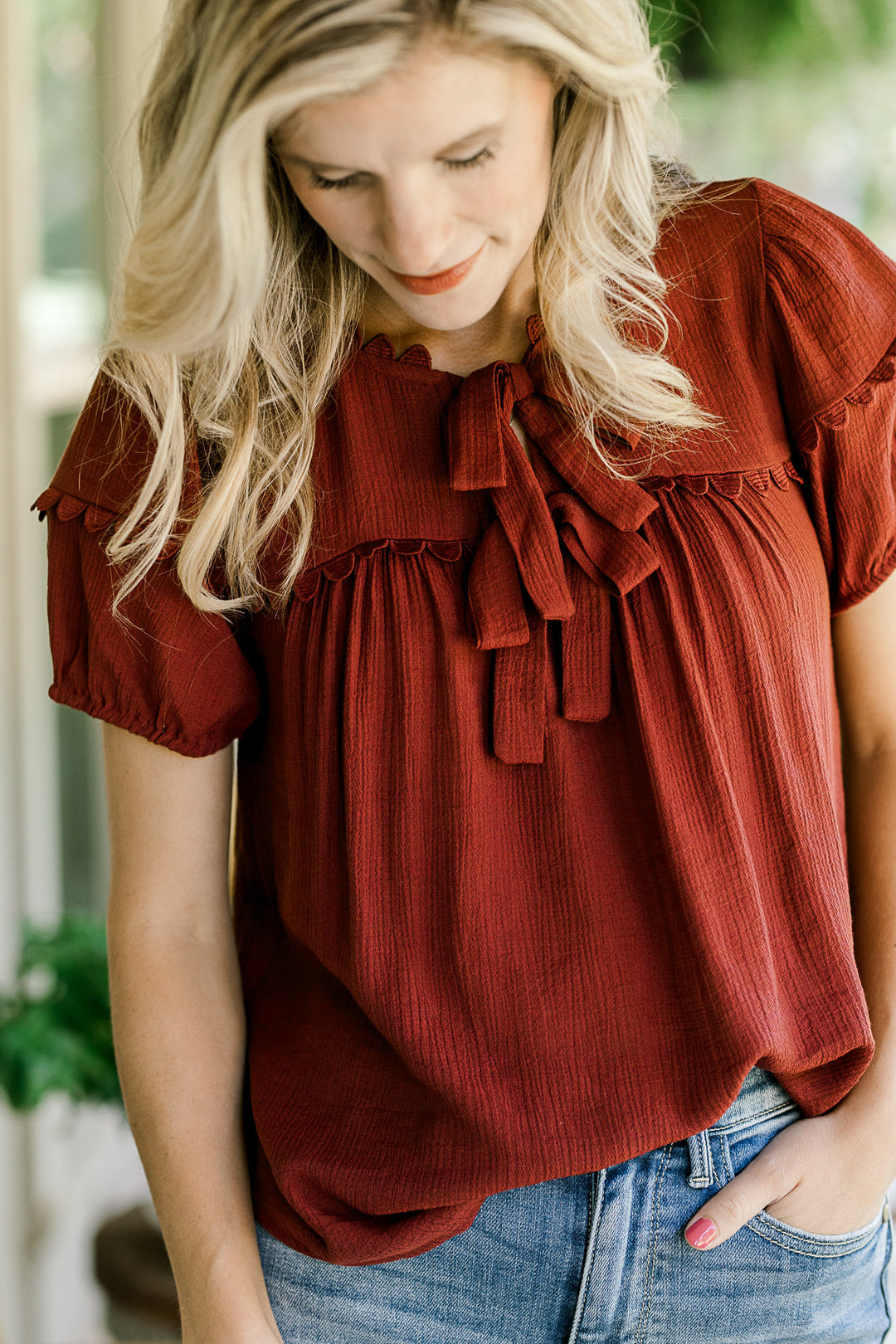 Model wearing a burgundy top with scalloped detailing, v-neck with tie and short sleeves. 