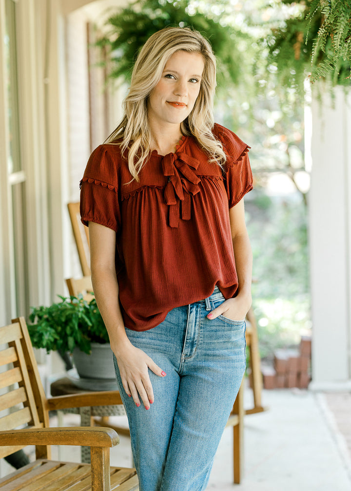 Model wearing jeans and a burgundy top with scalloped detailing, v-neck with tie and short sleeves. 