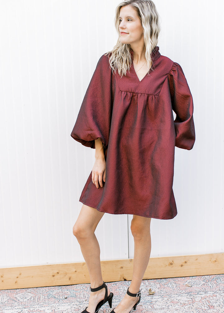 Model wearing heals and a burgundy dress with a v-neck, 3/4 bubble sleeves and ruffle neck.