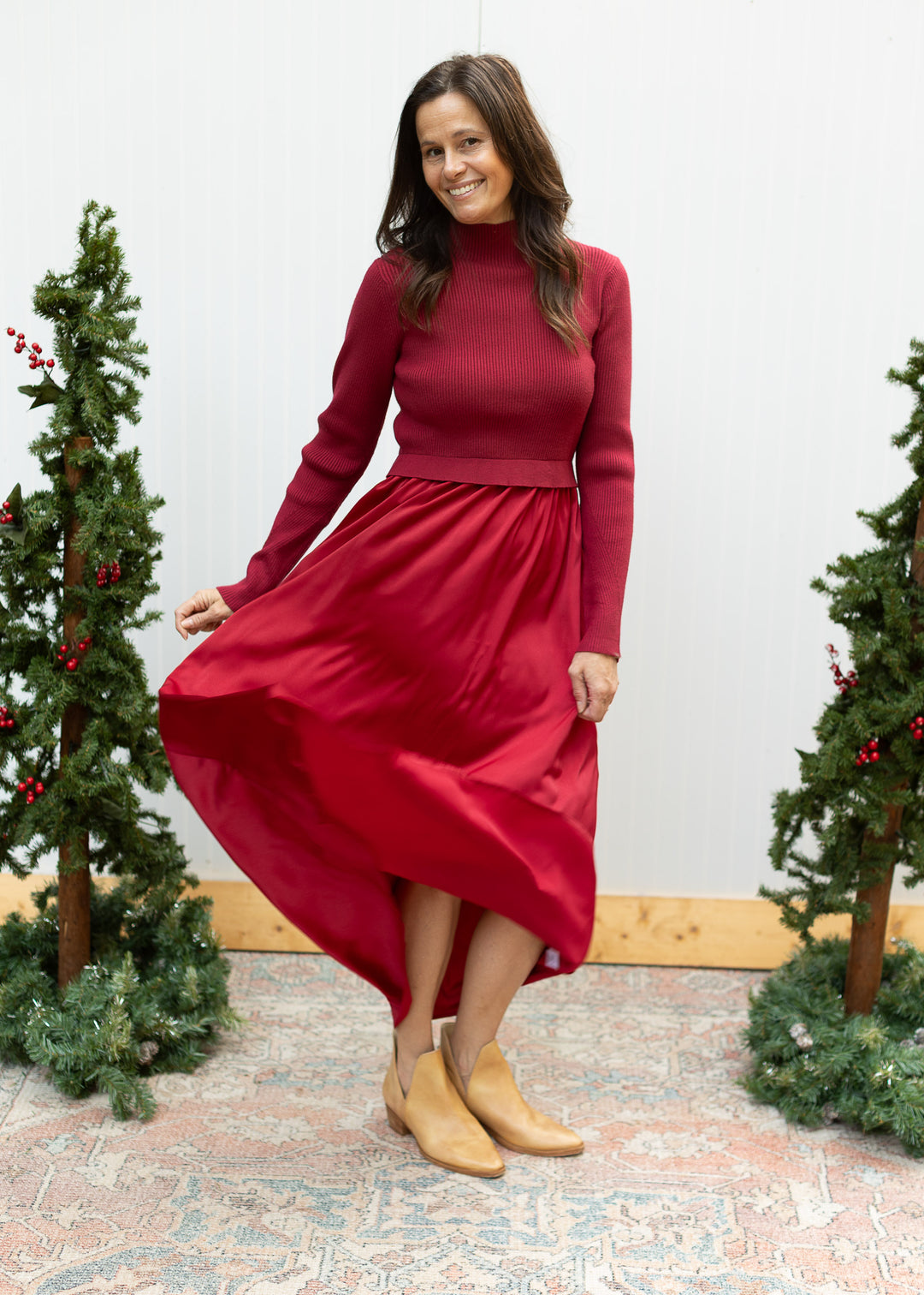 Model lifting the flowing satin skirt of a burgundy maxi dress with a ribbed sweater bodice and long sleeves.