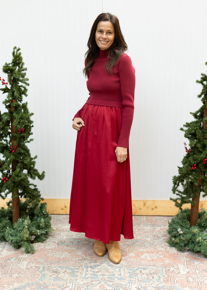 Full-length burgundy sweater dress with mock neck, ribbed long-sleeve bodice and satin skirt, styled with tan boots.