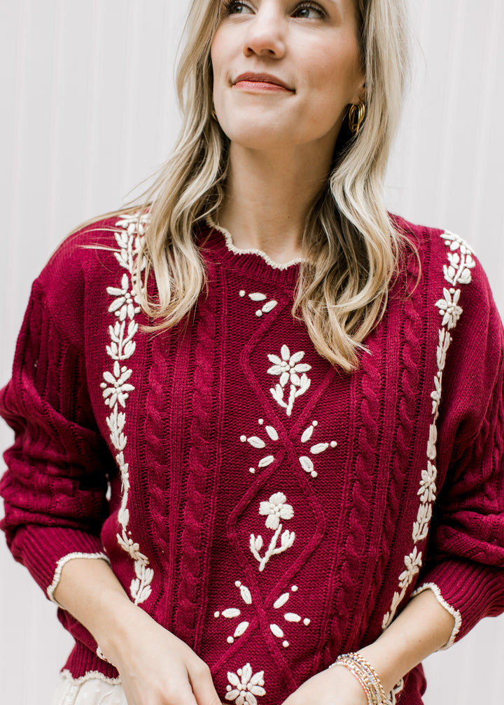 Close up of cream floral and scalloping at neck and sleeves of a burgundy long sleeve sweater. 