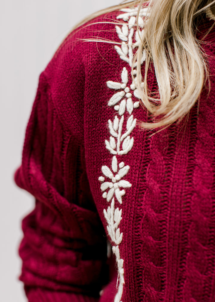 Close up of cream floral detail on a burgundy cable knit sweater with long sleeves. 