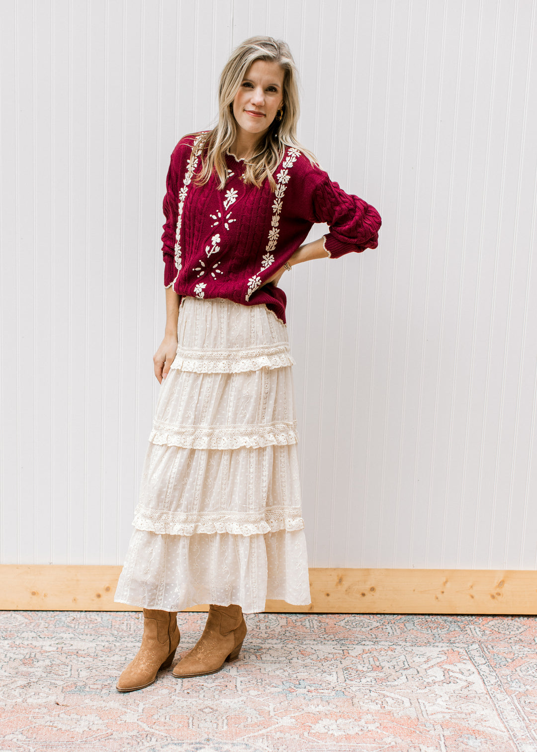 Model wearing a cream skirt, boots and a burgundy cable knit sweater with cream floral. 