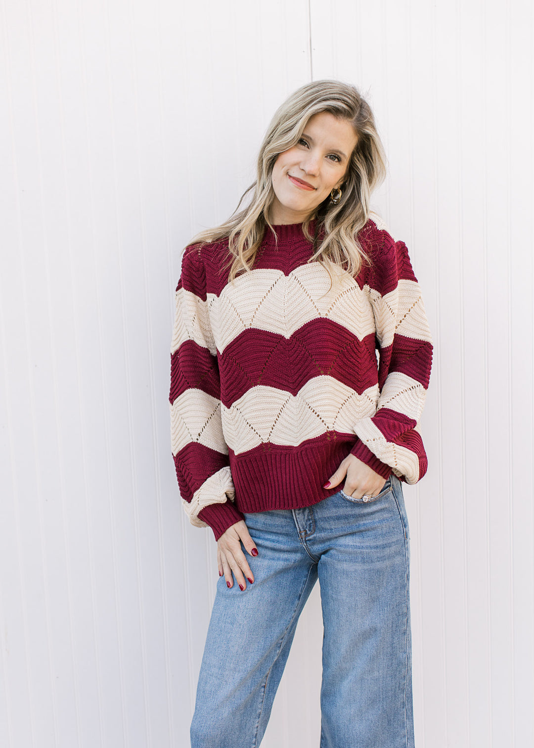 Model wearing jeans and a long sleeve cream and burgundy sweater with scalloped stripes. 