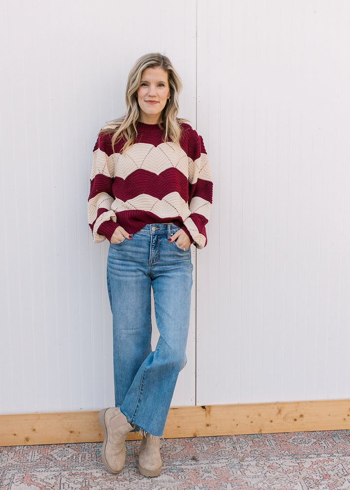 Model wearing jeans, boots and a long sleeve cream and burgundy sweater with scalloped stripes. 