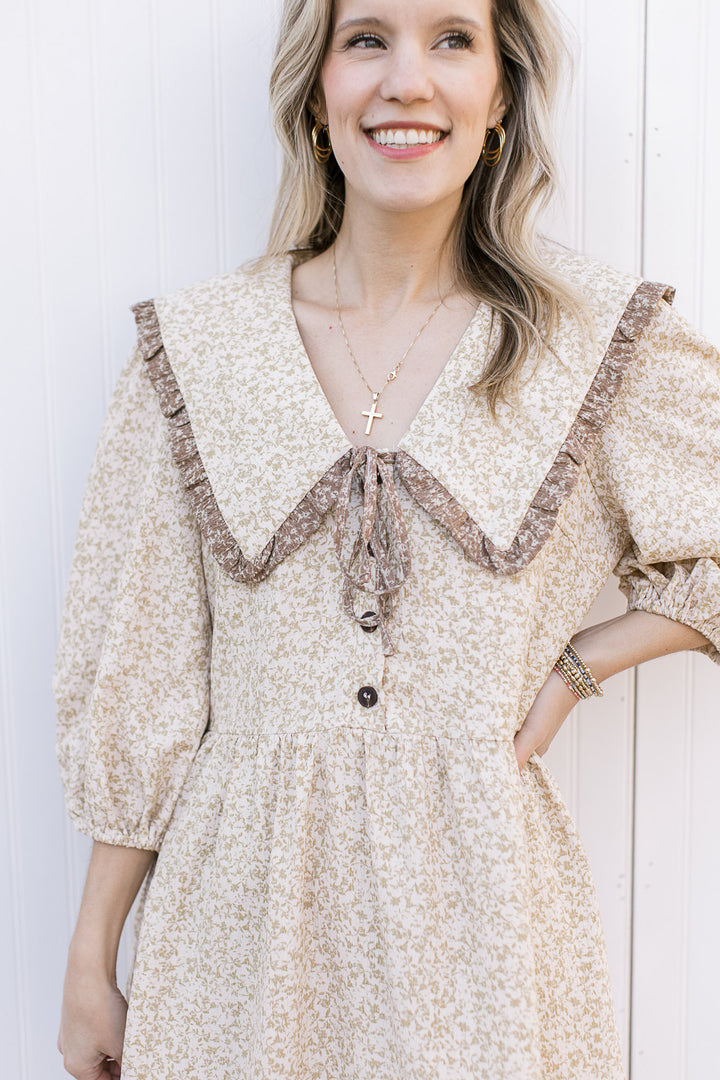 Close up of buttons and contrast taupe ruffle on wide v-neck collar of a cream dress with floral. 