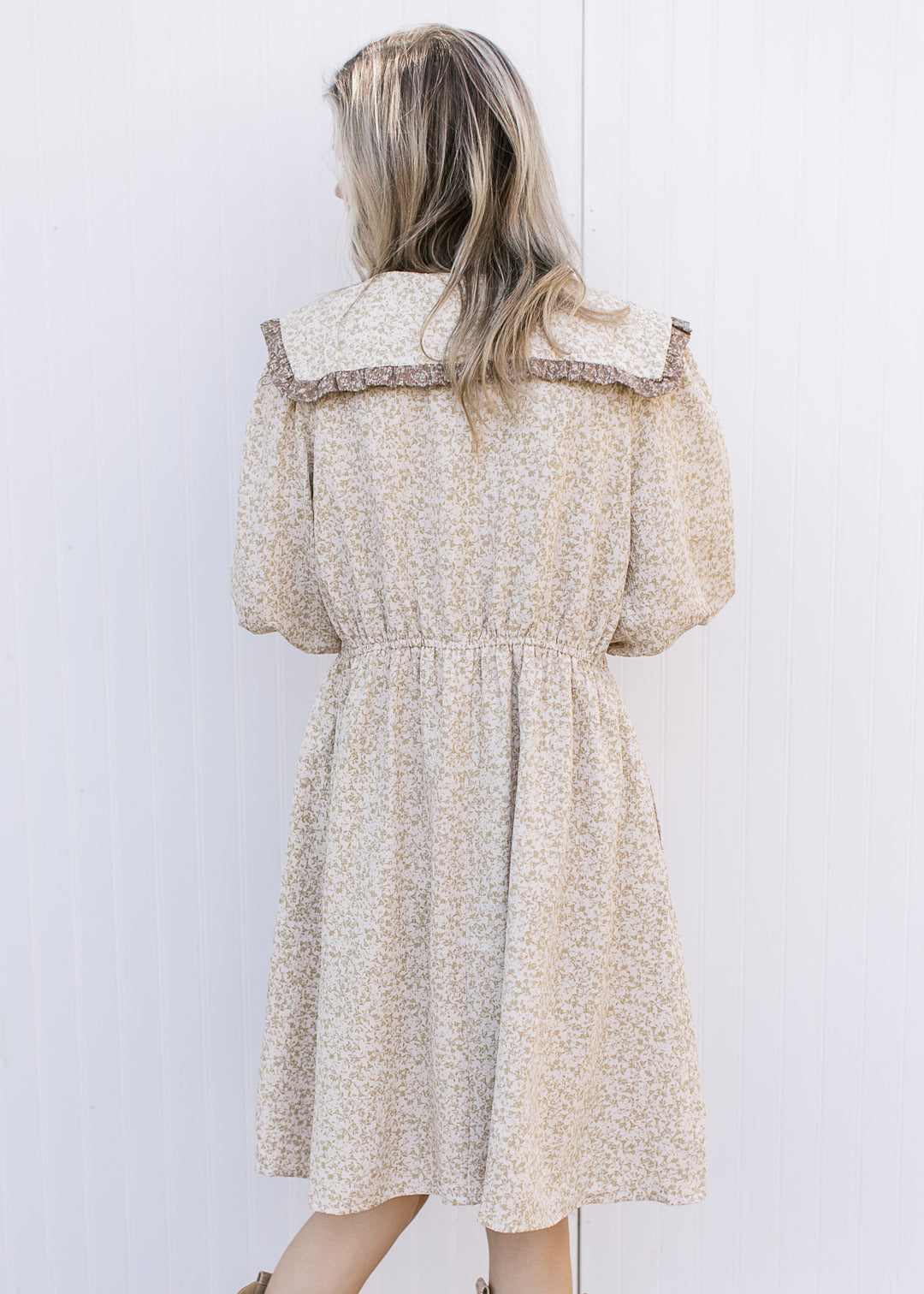 Back view of Model wearing a cream dress with taupe floral, 3/4 sleeves and a wide v-neck collar.