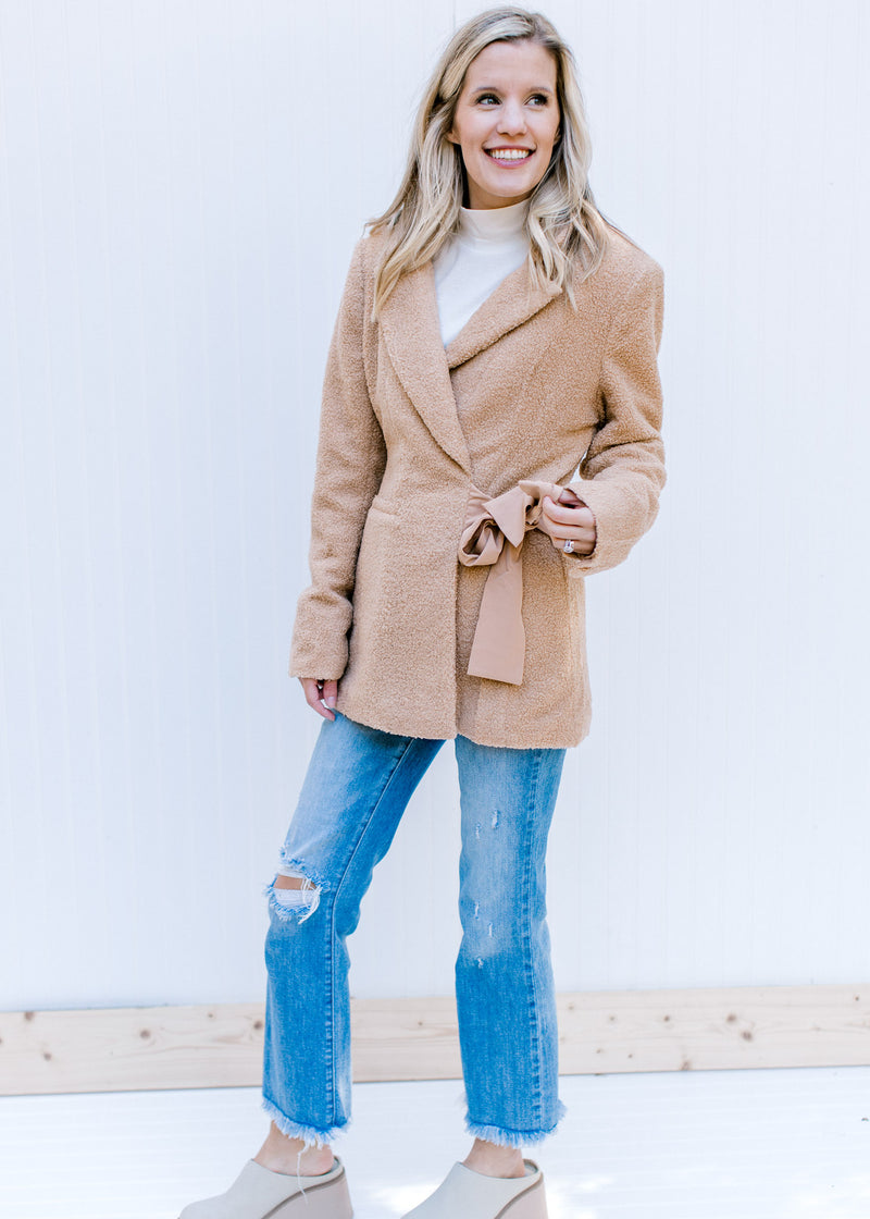 Model wearing jeans, mules and a white top with a camel long sleeve coat with  a side tie closure. 