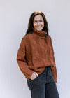 Model wearing a rust/camel colored sweater with a chunky cable knit, turtleneck and long sleeves. 