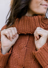 Close up of turtleneck and chunky cable knit on a rust/camel colored long sleeve sweater. 
