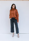 Model wearing jeans and a rust/camel colored sweater with a chunky cable knit and a turtleneck. 