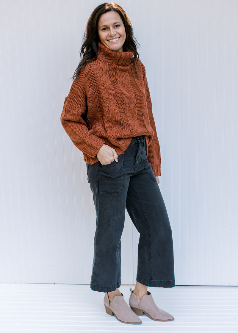Model wearing jeans, booties and a rust/camel colored sweater with a chunky cable knit design. 