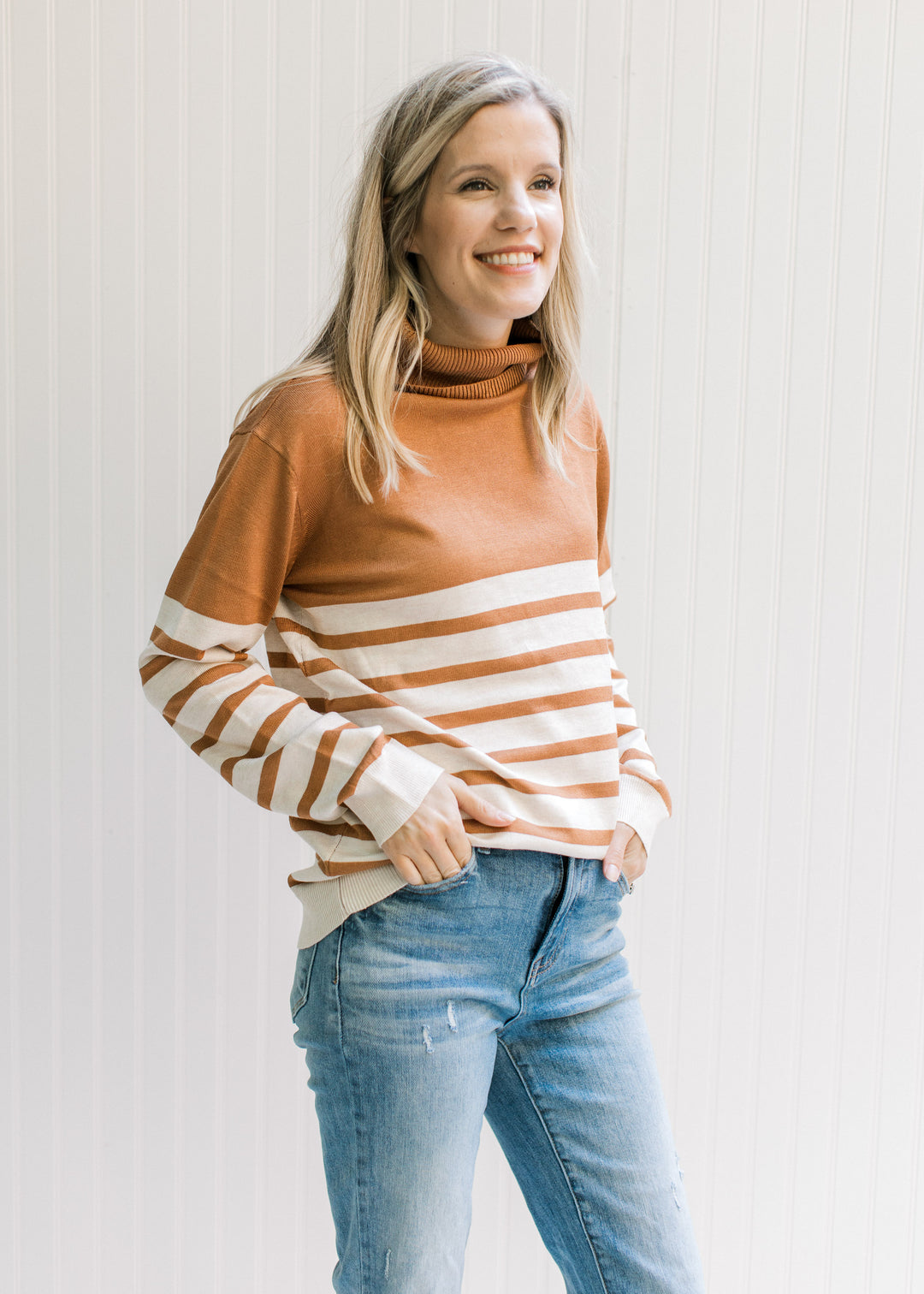 Model wearing jeans and a caramel and cream striped sweater with a turtleneck and long sleeves. 
