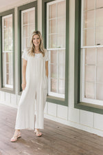 Model waering a white top and sandals with a sand jumpsuit with wide legs.