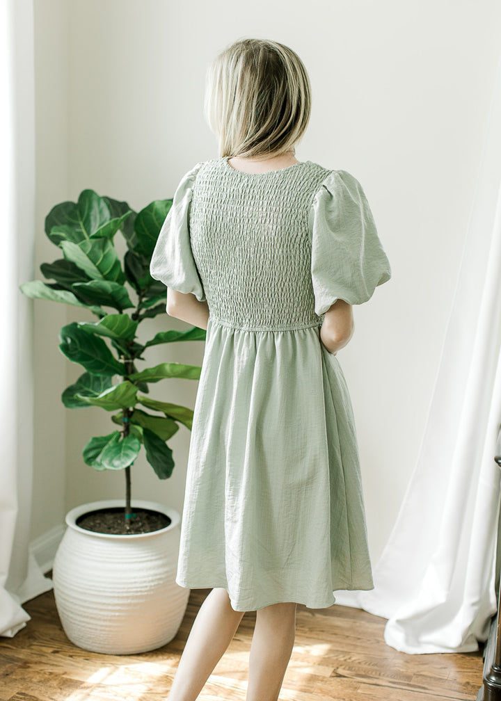 Back view of a short sleeve, knee length soft green dress with a smocked bodice and pockets.