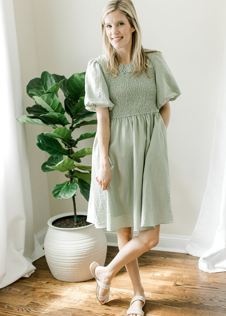 Model wearing sandals and a soft sage dress with a round neck, smocked bodice and short sleeves.