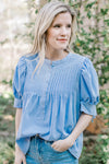 Model wearing a soft blue woven top with micro pleats at the bodice and short sleeves with a tie.