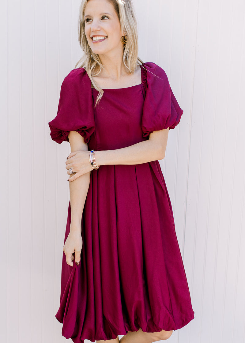 Model wearing a deep cranberry dress with a pleated waist, bubble short sleeves and hem. 