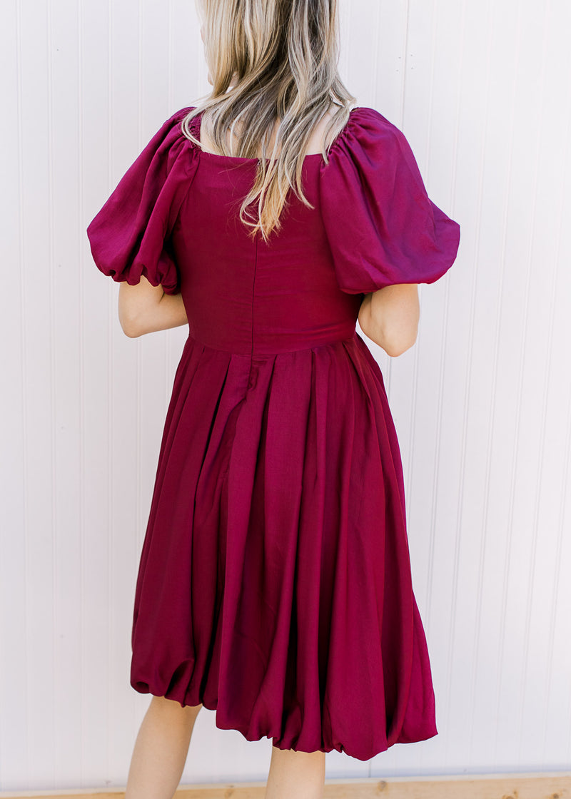 Back view of Model wearing a deep cranberry dress with bubble short sleeves and hem. 