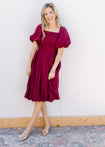 Model wearing gold flats and a deep cranberry dress with bubble short sleeves and hem. 