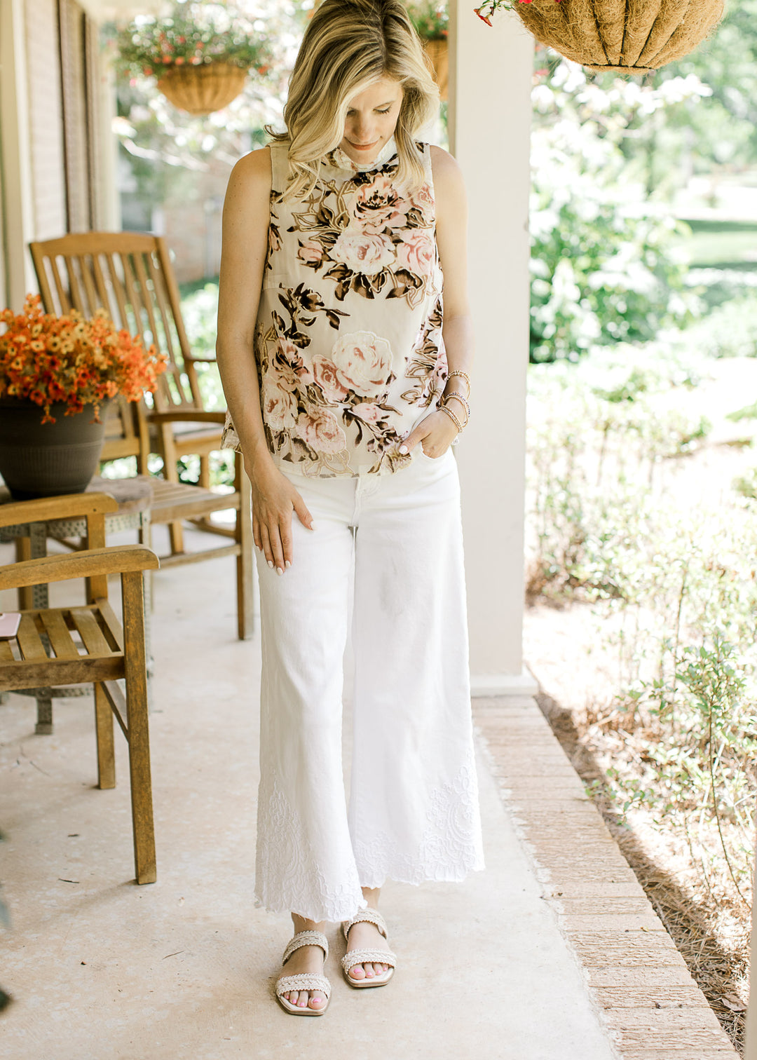Model wearing a white pants, sandals and a sleeveless oatmeal top with velvet pink and brown floral.