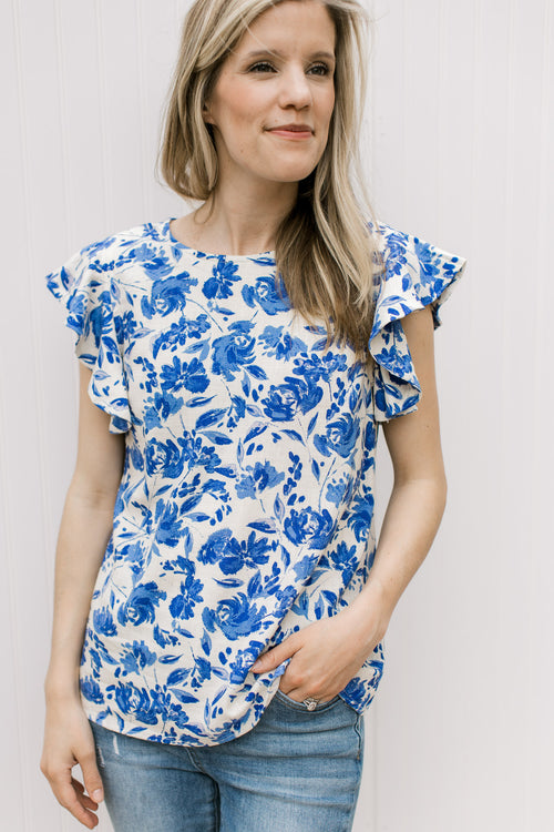 Model wearing a cream top with bright blue floral pattern and ruffle cap sleeves.