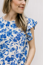 Close up of ruffle cap sleeve on a cream top with a bright blue floral pattern. 