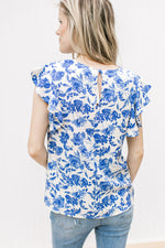 Back view of Model wearing a cream top with bright blue floral, cap sleeves and a keyhole closure.
